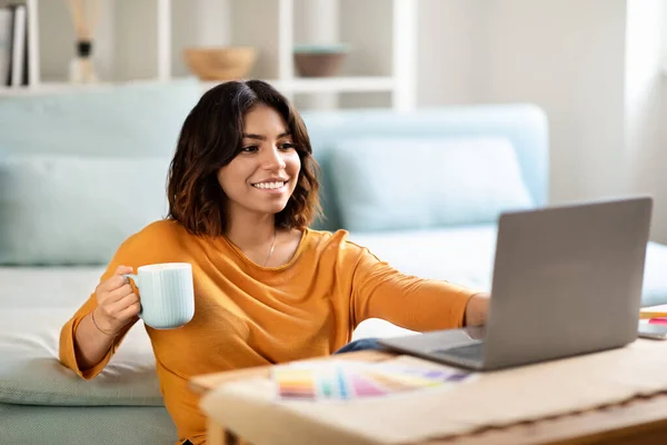 Distance Work. Smiling Arab Female Freelancer Drinking Coffee And Working With Laptop At Home, Happy Young Middle Eastern Woman Browsing Internet On Computer And Enjoying Hot Drink, Copy Space