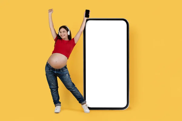 素晴らしいアプリだ 黄色の背景に白い画面で大きな空白のスマートフォンの近くに立っているヘッドフォンで喜び妊娠中の女性 幸せな期待の女性音楽ストリーミングアプリケーションをお勧めします モックアップ — ストック写真