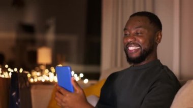 Mesafeli ilişki. Genç, mutlu, Afro-Amerikalı bir adam kız arkadaşıyla internetten sohbet ediyor, akıllı telefona el sallıyor, akşamları evde dinleniyor, çekim takip ediyor, yavaş çekim, serbest alan.