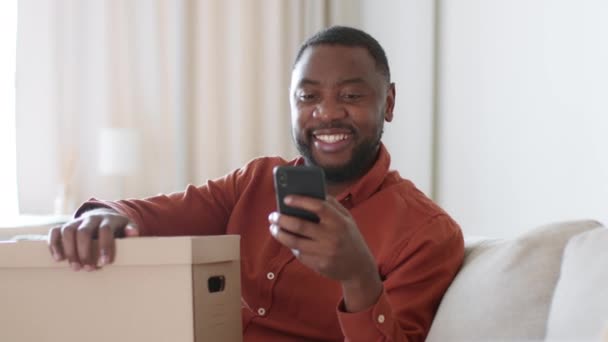 网上购物应用程序 快乐的年轻非洲裔美国人顾客在智能手机上打字 带着送货箱坐在家里 跟踪镜头 慢动作 自由自在 — 图库视频影像