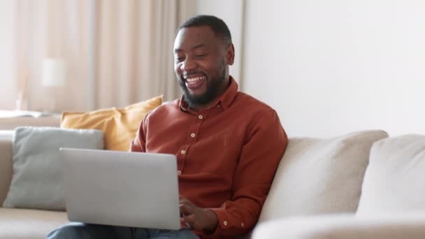 素晴らしいストリーミングサービス 若い幸せなアフリカ系アメリカ人男性はノートパソコンでデジタルテレビを見て笑って 自宅でお気に入りのショーを楽しんで 撮影を追跡し スローモーション 空のスペース — ストック動画