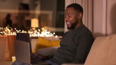 Çevrimiçi toplantı. Genç, Afro-Amerikan bir adamın laptopta arkadaşlarıyla görüntülü konuşması, el sallaması ve gülmesi, akşam evde oturup çekim yapması, yavaş çekim, serbest alan.