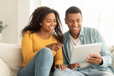 Neşeli Afrikalı Afro-Amerikan karısı koca tabletine parmağını gösteriyor, oturma odasının içinde görüntülü görüşme yapıyor. Sohbet teknolojisi, uzaktan buluşma uygulaması ve evde internette sörf.