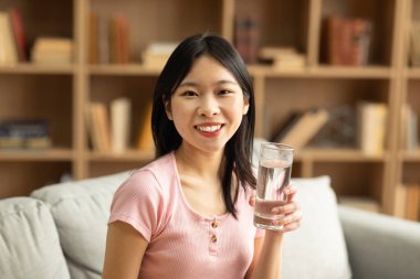 Oturma odasında kanepede oturmuş kameraya gülümserken elinde bir bardak temiz su tutan mutlu Asyalı kadın. Güzel bayan sağlığına ve sağlığına dikkat ediyor.