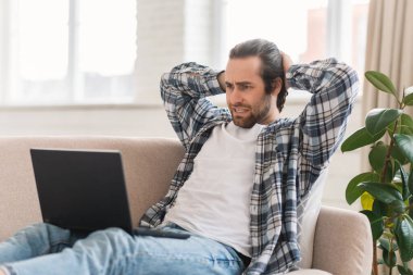 Neşeli, genç, beyaz bir adam kanepede bilgisayar başında oturur, video izler, dinlenir, oturma odasında tek başına vakit geçirir. Blog, aygıt, reklam ve teklif, video çağrısı ve sosyal uzaklık ile rahatla