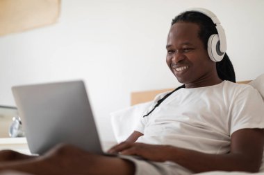 Çevrimiçi Eğitim. Genç siyahi adam evde yatağında yatarken dizüstü bilgisayarla çalışıyor kablosuz kulaklık takmış, uzaktan öğrenme için bilgisayar kullanarak gülümseyen bir Afrikalı Amerikalı.