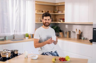 Gülümseyen yakışıklı, bin yıllık beyaz sakallı, ev kıyafetleri ve kahve fincanı olan adam günaydın, mutfağın içinde tek başına kahvaltı yapıyor. Hafta sonları, sağlık hizmetleri ve boş zamanlarında ruh hali