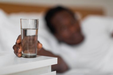 Yatağın başucundaki masadan bir bardak su alan susamış siyah adam sabah uyandıktan sonra sağlıklı içeceğe uzanan Afrika kökenli genç erkek seçici bir şekilde boş alana odaklanıyor.