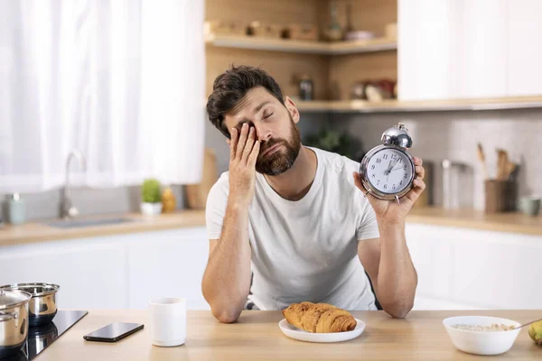 目覚まし時計付きのひげを持つ不幸な疲れ眠い千年紀の白人男性は テーブルで彼の目をこすり キッチンのインテリアだけで朝食を持っています 睡眠不足 早起き 働く時間 — ストック写真