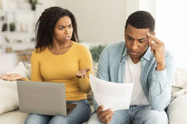 Angry unhappy millennial african american lady quarreling at guy over debts and bills with laptop in living room interior. People emotions, problems with finances, bankruptcy, taxes and crisis at home