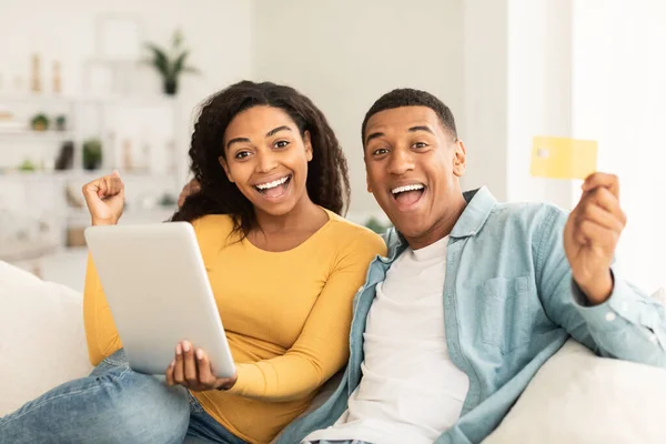 Happy surprised millennial african american couple use tablet and credit card for shopping online, make victory and success hand gesture in living room interior. Win emotions, sale and huge cashback