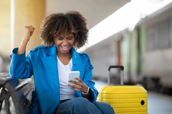 快乐的黑人女人坐在火车站的长椅上用智能手机庆祝成功 快乐的非洲裔美国女人玩网络游戏 一边等火车一边玩乐 自由自在 — 图库照片