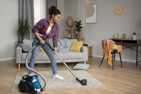 Lifestyle and work at home. Smiling millennial african american lady cleaning dust with vacuum cleaner in living room interior, full length. Household chores, health care, hygiene and cleaning service