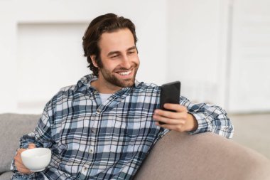 Kafkasyalı genç bir erkeğin kanepede oturup, bir fincan kahveden ve akıllı telefondan sohbetten hoşlanması ve odanın içinde iyi haberler okuması iyi bir şey. Komik videolar izleyin, sosyal medyada mesajlaşma ve boş zamanlarda blog için uygulama