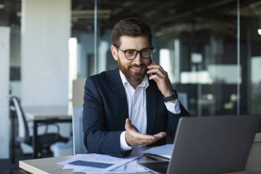 Cep telefonuyla konuşan pozitif iş adamı, modern ofiste dizüstü bilgisayarla iş yerinde oturan, kopyalama alanı. Erkek çalışan telefonla konuşuyor. İş iletişimi