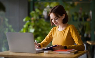 Masada oturan genç bir kadın, not defterine not alıyor, webinar ya da eğitime katılıyor, kafedeki modern bilgisayar ve kulaklıkları kullanıyor. İnternette kız öğrenci öğrenimi, eğitim kursuna katılma, kopyalama alanı