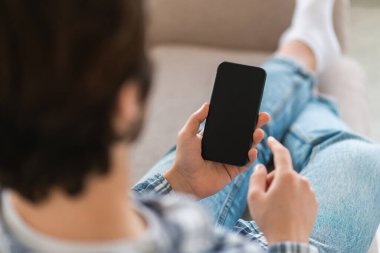 Kırpılmış genç beyaz adam kanepeye oturur, boş ekranla akıllı telefonda sohbet eder, internette sörf yapar, oda içinde haber okur. Komik videolar izleyin, sosyal medyada mesajlaşma, uygulama ve blog arama
