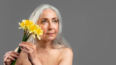 Beyaz saçlı, sarı çiçekli, ciddi bir beyaz kadın gri arka planda ve stüdyoda izole edilmiş boş alana bakıyor. Güzellik ve cilt bakımı, yaşlanma karşıtı tedavi, spa prosedürü, reklam ve sunum