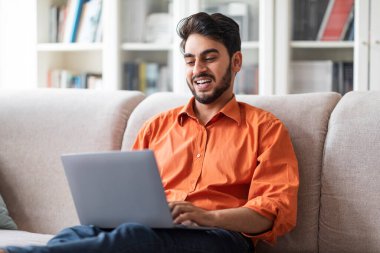 Mutlu, sakallı, serbest çalışan genç, orta doğulu bir adam oturma odasında oturuyor, dizüstü bilgisayar, programlama, kopyalama alanı kullanıyor. Uzak görev konsepti