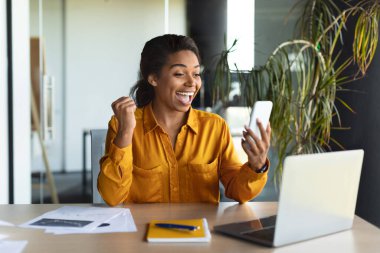Heyecanlı siyah kadın akıllı telefondan güzel mesajlar okuyor, modern ofis içinde dizüstü bilgisayarla çalışıyor. Çevrimiçi uygulama ile harika haberler, başarılı iş, geliştirme, planlama ve finansal stratejiler