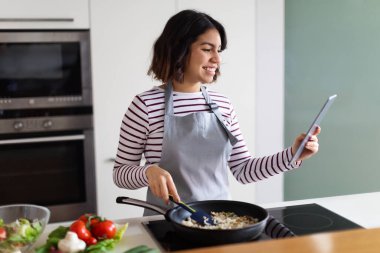 Neşeli, güzel, genç, esmer, orta doğulu kadın evde yemek yapmayı, yemek tarifini, internette yemek blogunu, modern dijital blogu kullanarak mutfakta sağlıklı yemek hazırlıyor, uzayı kopyalıyor.