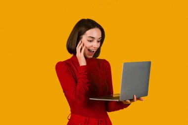 Güzel online teklif. Çevrimiçi kırmızı elbiseli genç bayan, elinde modern laptop, bilgisayar ekranına bakıyor ve bağırıyor, turuncu arka planda poz veriyor, boşluğu kopyalıyor