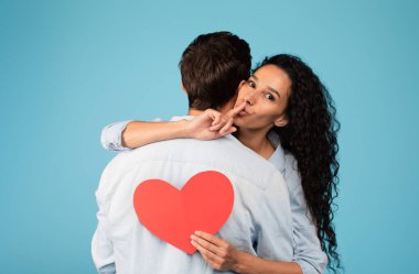 Neşeli genç Arap kadın Avrupalı kocaya sarılıyor, yüreğini tutuyor, mavi arka planda parmak işareti var. Gizli jest, sürpriz, hediye, aşk ve ilişki, sevgililer günü, yıldönümü kutlaması