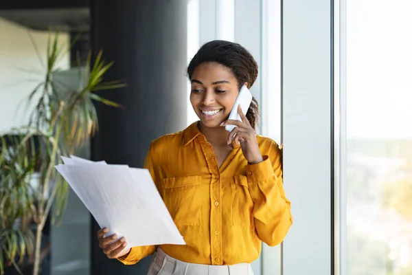 Corporate communication. Smiling african american female manager holding financial documents and talking on cellphone, standing near window in office interior
