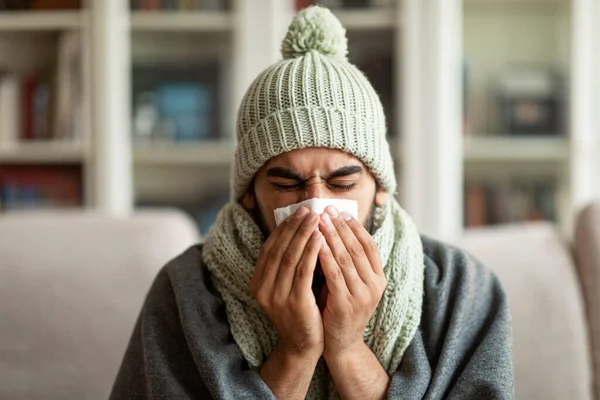 Cold, flu, seasonal disease. Unhappy young arab man feeling bad, wearing knitted ice hat, sitting on sofa covered in warm blanket, blowing his nose at home, living room interior, closeup, copy space