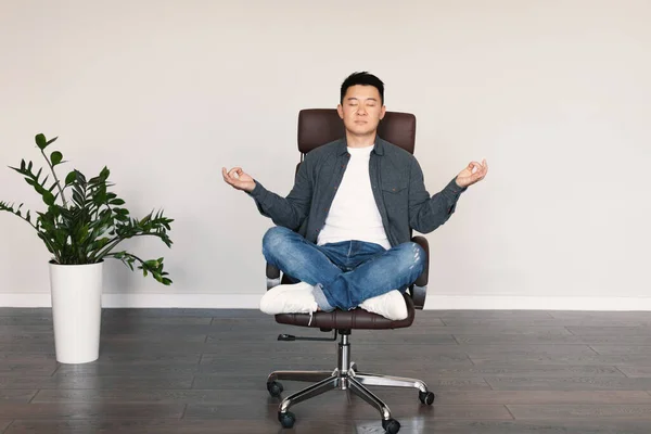 Serious calm adult asian male with closed eyes sits in armchair, meditates, rests from work, enjoys silence and free time in living room interior with white wall. Relax at home, break, lifestyle