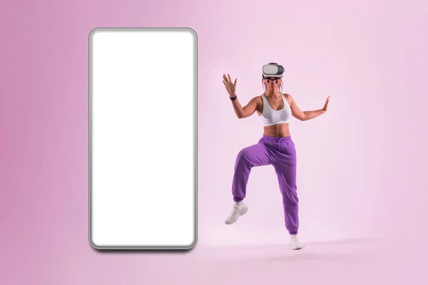 Dance moves in metaverse. Young woman dancing in virtual reality headset near huge smartphone with blank screen, pink background, mockup. Black lady having fun while exploring 3D technology.