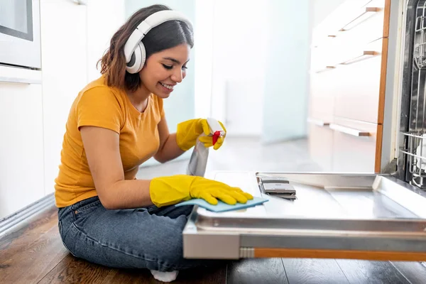 Smiling Middle Eastern Female Wiping Dishwasher Machine Surface While Doing Cleaning In Kitchen, Happy Young Arab Woman Wearing Wireless Headphones Using Rag And Sprayer While Tidying Home