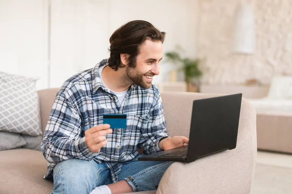 Glad young caucasian guy shopaholic sits on sofa with computer and credit card, shopping online, checks financial account, ordering goods in room interior. Application banking, sale, ad and offer
