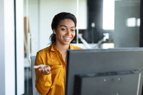 Positive black female entrepreneur gesticulating at computer webcamera, working and having video call in office interior. Business meeting and modern technology