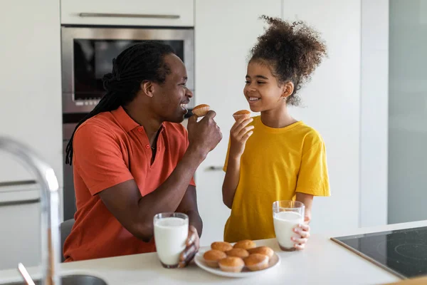キッチンでお父さんと一緒にお菓子を食べるかわいいプリティーブラックガールの肖像 幸せなアフリカ系アメリカ人の父と娘自宅で一緒に一口を持ちながらおいしいマフィンと牛乳を飲みながら楽しむ — ストック写真