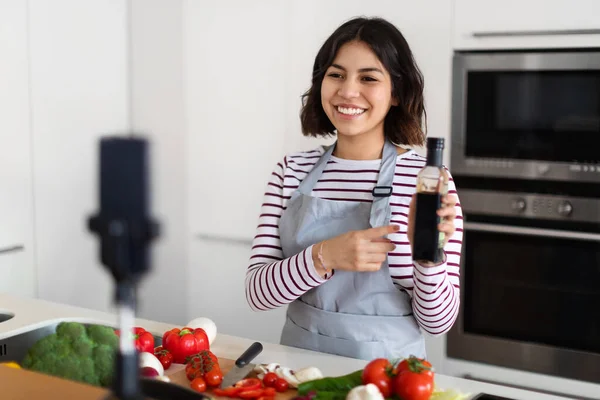 Food blogging. Positive pretty millennial brunette woman recording food vlog, female blogger cooking at home, showing bottle with sauce or oil at smartphone camera and smiling, kitchen interior
