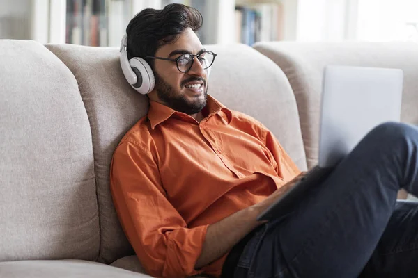 Happy handsome young middle eastern man wearing eyeglasses and casual outfit reclining on sofa, using wireless headphones, looking at computer screen, smiling, gaming, home interior, copy space