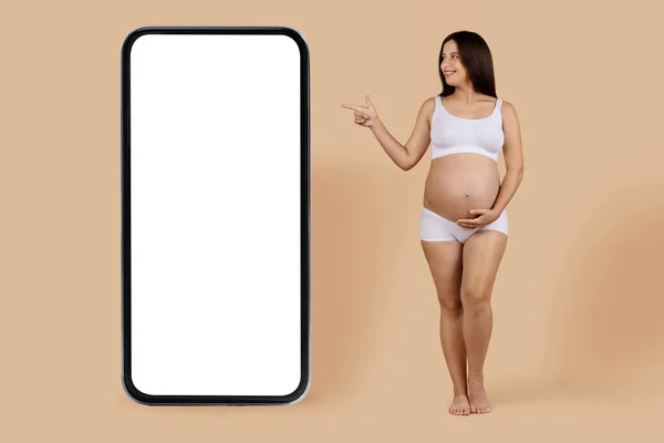 オンライン広告 白い画面で大きな空白のスマートフォンで脇を指して幸せな若い妊娠中の女性 下着で期待される女性を笑顔コピースペースを実証 ベージュの背景に立って モックアップ — ストック写真