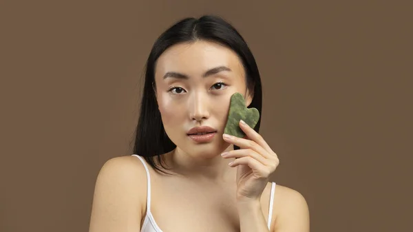 Korean lady massaging face by gua sha quartz stone scraper for skincare, posing over brown background, panorama. Woman using natural massager. Face massage, women beauty trends.