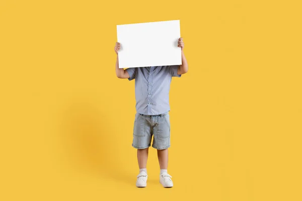 Kids ad. Full length shot of unrecognizable little boy covering his face with blank sheet of paper, orange studio background, empty space