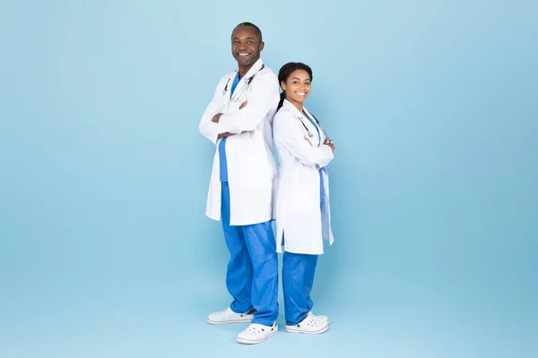 Mature african american doc man and woman posing with folded arms, experienced doctors wearing lab coats, standing over blue studio background, full body length