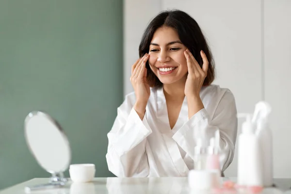 Skin care routine. Happy attractive young middle eastern woman in white silk bathrobe using eyes cream at home, beautiful lady test newest organic beauty product, sitting at vanity table, copy space