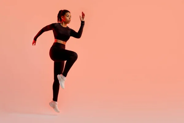Sporty black lady jumping, having cardio training, wearing black fitwear, exercising over neon peach studio background, free space. Fitness workout concept. Full length