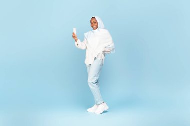 Cep telefonu kullanan Afrikalı Amerikalı Müslüman kadın, mavi arka planda poz veriyor, stüdyo çekimi, tam boy. Başörtülü kadın akıllı telefon tutuyor, yeni mobil uygulama kullanıyor