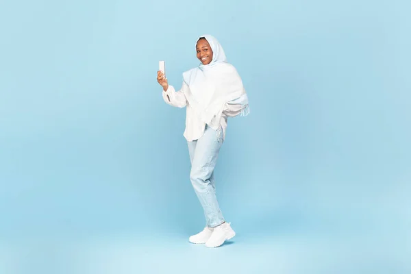 African american muslim woman using cellphone, posing on blue background, studio shot, full length. Lady in hijab holding smartphone, using new mobile application