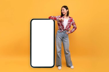 Beyaz ekranlı, gülümseyen, sarı stüdyo arka planlı, büyük akıllı telefona yaslanan mutlu Japon kadın. Mobil uygulama, web sitesi, reklam tasarımınız için şablon