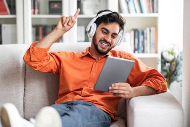 Heyecanlı yakışıklı, sakallı, orta doğulu genç adam evdeki koltukta dinleniyor, yeni dijital tablet ve kablosuz kulaklıklar kullanıyor, arkadaşlarıyla online oyunlar oynuyor, video izliyor, yer kopyalıyor.