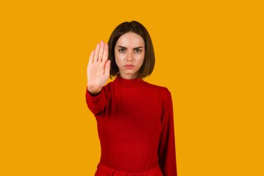 Ciddi bir şekilde kendine güvenen, kırmızı elbiseli esmer bir kadının portresi uzanmış elleriyle ayakta duran, dur hareketi gösteren bir kadın turuncu stüdyo arka planında izole edilmiş, kopyalama alanı