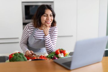 Heyecanlı, genç, esmer, orta doğulu bir kadın evde yemek pişirmekten zevk alıyor, internette yemek tarifi okuyor, internette yemek blogunu kontrol ediyor, mutfakta sağlıklı yemek hazırlarken dizüstü bilgisayar kullanıyor, uzay kopyaları yapıyor.