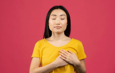 Sarı tişörtlü mutlu sakin genç Çinli kadın ellerini göğsüne, kalbine bastırıyor, pembe arka planda poz veriyor, stüdyo çekimi yapıyor. İnsanlar duygular, minnettarlık, teşekkür ve sevgi